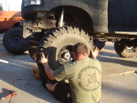 Man in green t-shirt working on a large off-road tire. Another orange vehicle is visible in the background