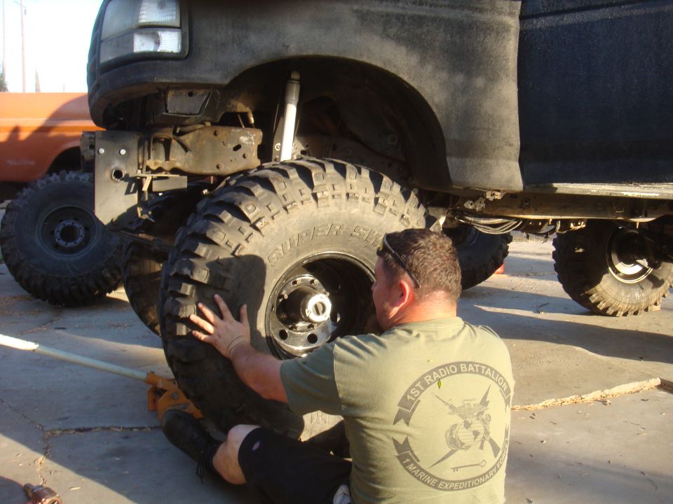 Man in green t-shirt with large tires and jack under dark truck