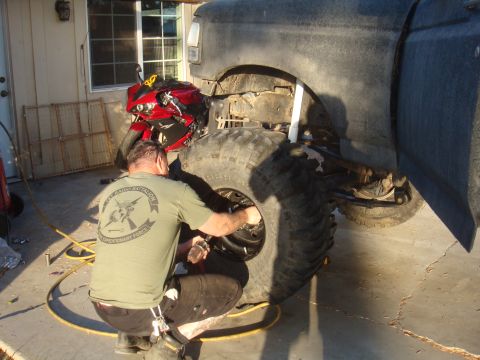 Man crouches to work on a massive tire mounted on a truck; red motorcycle parked nearby
