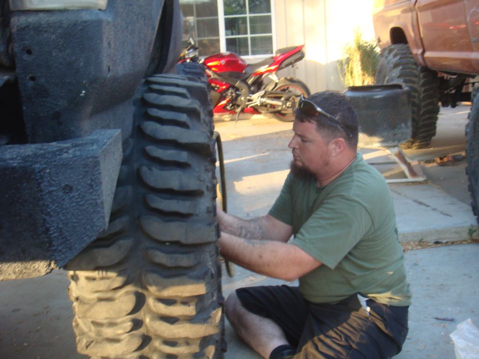 Man working on a large off-road tire, with a red motorcycle and a lifted truck in the background