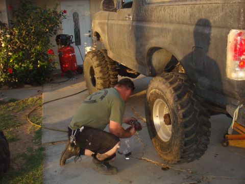 Man crouches to work on the rear axle of a lifted 1992 Ford Bronco. Large off-road tires are visible