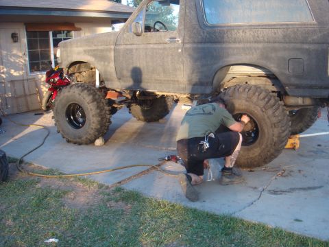 Man working on a lifted Ford Bronco with huge tires. A red motorcycle is parked nearby
