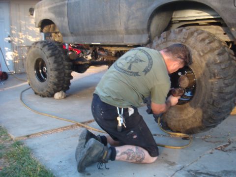 1992 Ford Bronco side view, man kneeling to work on a large tire, gray exterior, concrete driveway