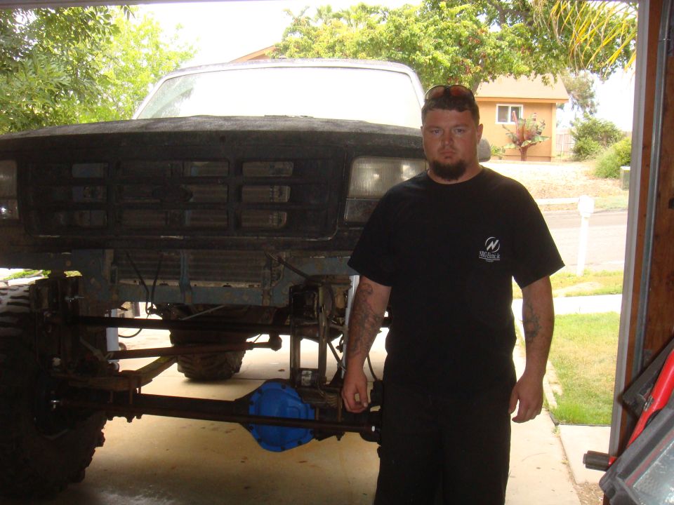 Man in black t-shirt stands before 1992 Ford Bronco front end and chassis. Truck is lifted with visible suspension and axle