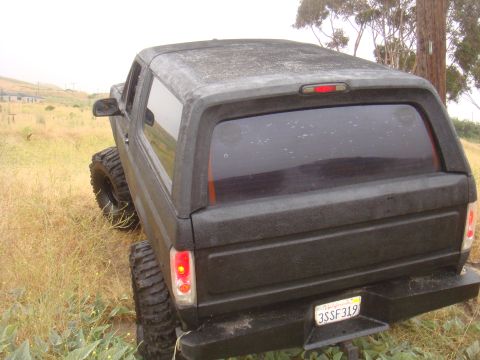 Black SUV with large off-road tires parked in a grassy field. Rear view shows tinted windows and a California