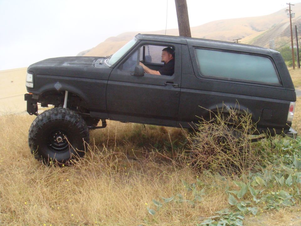 Black 1992 Ford Bronco with lifted suspension and large tires parked on dry grass, driver visible