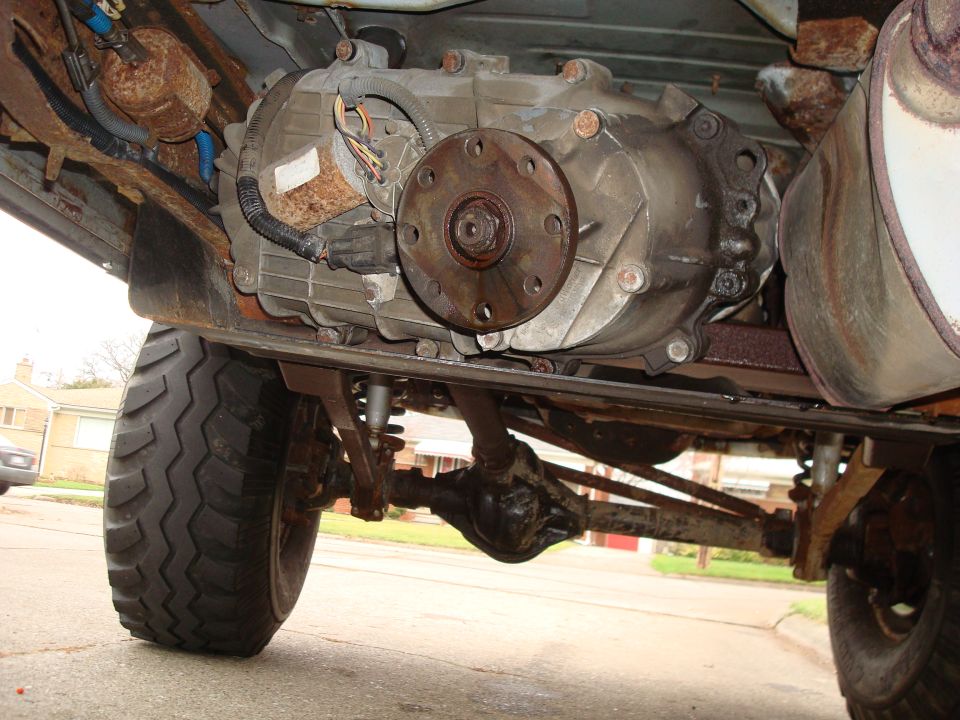 Transfer case and drivetrain of a vehicle, viewed from below. Rusty components and a large tire are visible