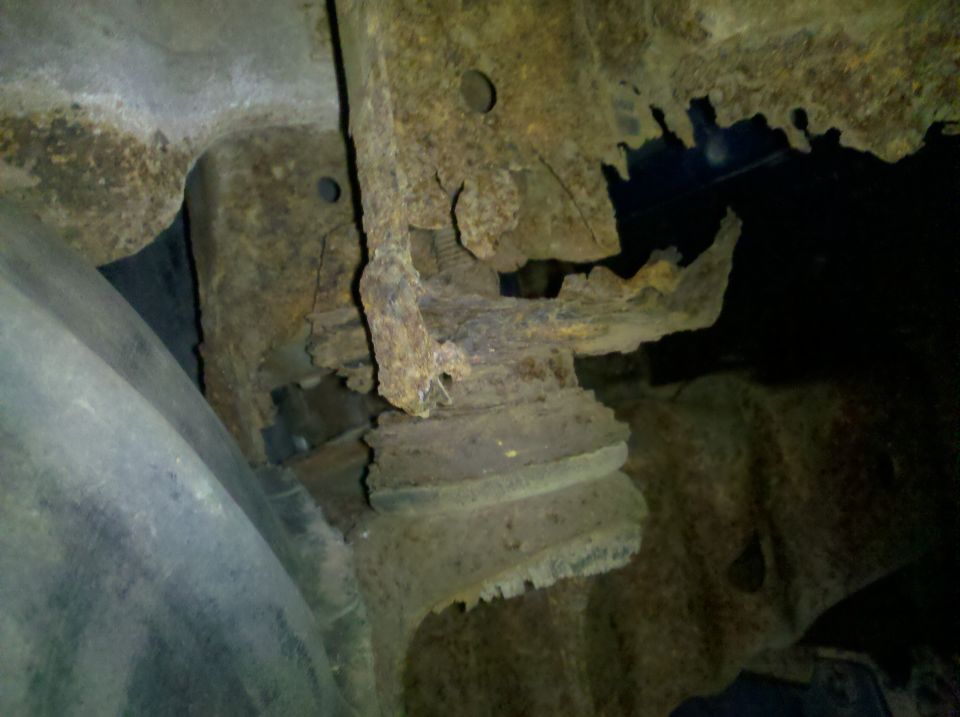 Severely rusted body mount and perforated frame metal underneath a vehicle, tire visible at left