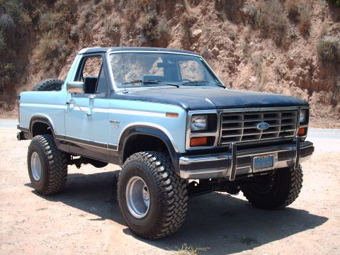 1986 Ford Bronco - This lifted Ford Bronco is parked on a dirt road with a rocky hillside in the background.
