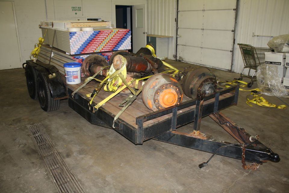 Trailer loaded with drywall, buckets, and large orange-hubbed axles. Yellow straps secure the load