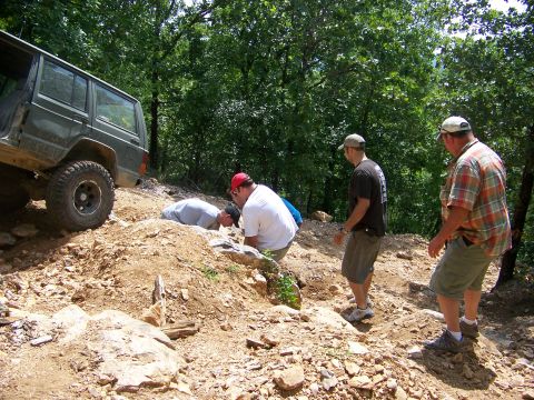 1992 Jeep YJ - Several people are gathered around the front wheel of a parked green SUV on a dirt path.