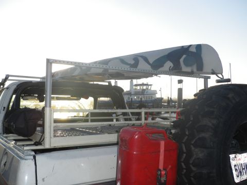 1991 Ford Bronco - Truck bed loaded with gear, including a spare tire and gas can, with a ferry in the distance.