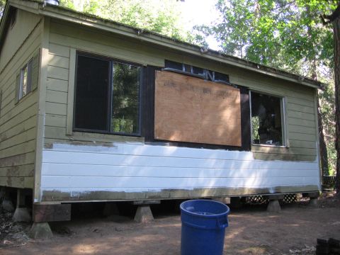 1989 Ford Bronco - A cabin sits on blocks with a boarded-up window and a blue trash can in front.