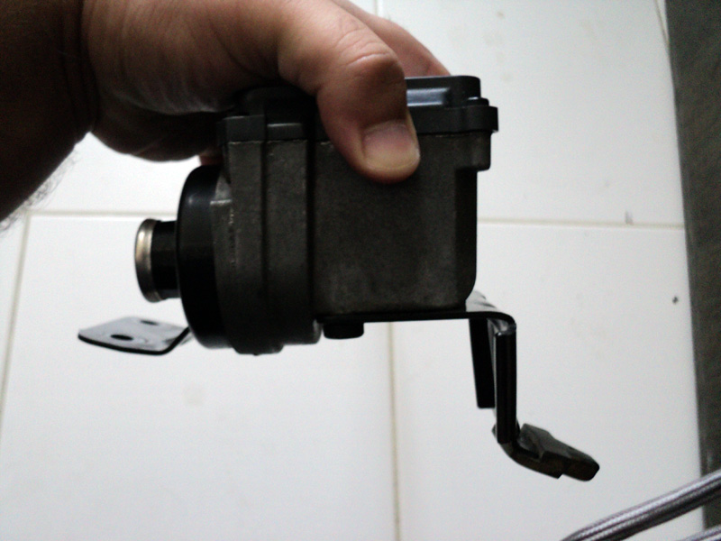 Hand holding a removed cast aluminum fluid reservoir with black cap and steel mounting bracket