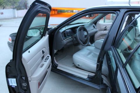 2007 Ford Crown Victoria - Driver's side interior of a black Ford sedan shot through the open front door, showing the gray dash, four-spoke Ford steering wheel with a…