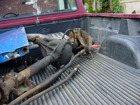 1992 Ford Bronco - Greasy salvaged driveline parts, a differential housing with axle tube, brake lines, a rusty steering box and a blue painted panel, loaded…