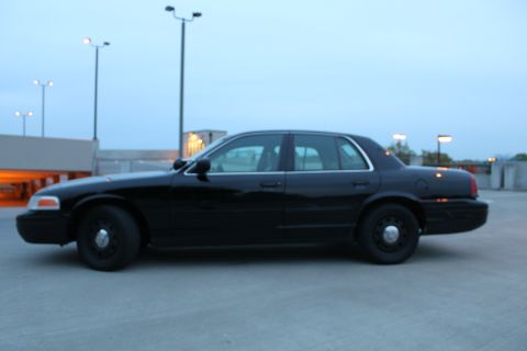 2007 Ford Crown Victoria - Driver-side profile of a black four-door full-size sedan parked on an open rooftop parking deck at dusk under light poles. It rides on plai…