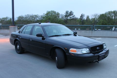 2007 Ford Crown Victoria - Three-quarter front view of a black Ford Crown Victoria police-package sedan on a concrete parking deck at dusk, with blacked-out grille, A…
