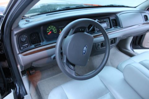 2007 Ford Crown Victoria - Driver's-side cockpit view through the open door of a Ford sedan, showing the four-spoke steering wheel with blue Ford oval airbag cover, g…