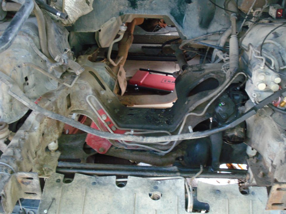 View into vehicle's engine bay, showing frame and some engine components. A red Craftsman jack is visible