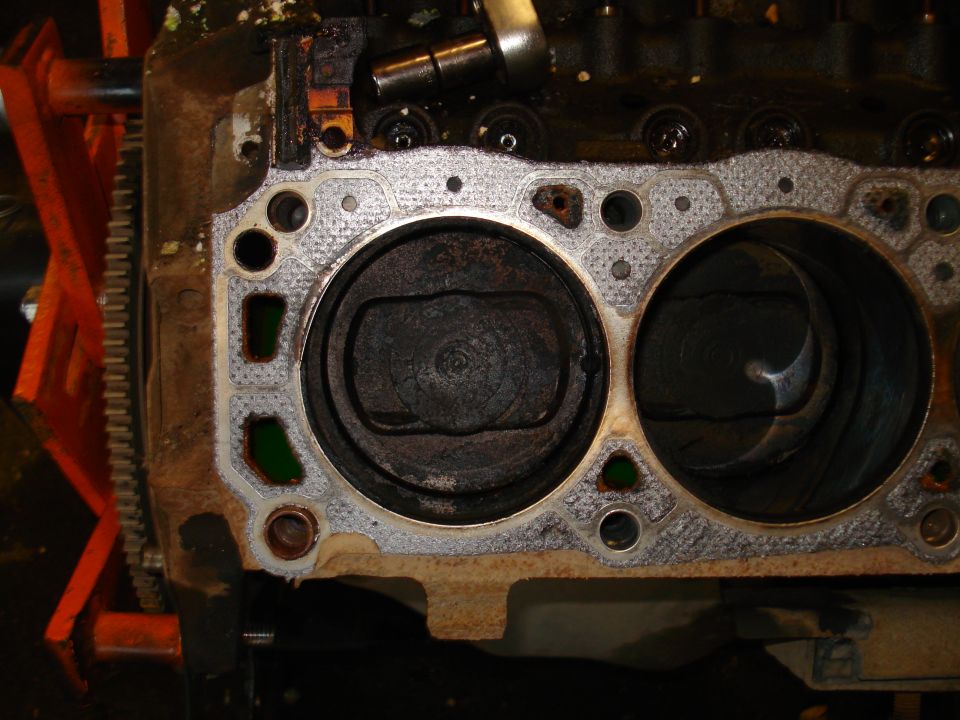 Engine block with two cylinders, pistons visible. Gasket is corroded. Orange stand supports the engine
