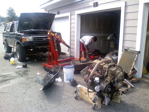 1990 Ford Bronco - Driveway engine-swap scene: a lifted black Ford full-size SUV sits with its hood up and engine pulled, a red hydraulic engine hoist parked…