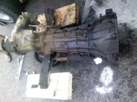 1990 Ford Bronco - Transmission assembly, likely removed from a vehicle, rests on wooden blocks on asphalt.