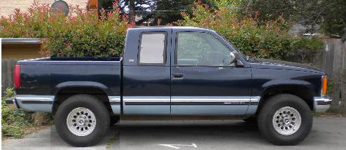 Blue and silver two-tone extended-cab pickup parked in profile by a wood fence