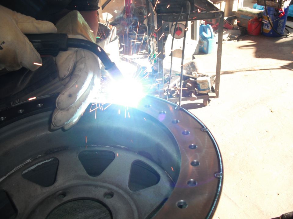 Person welding a metal wheel rim with many bolt holes around the edge
