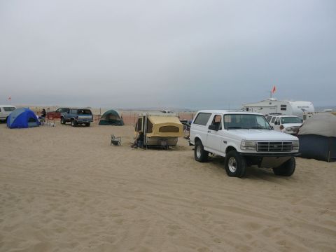 1988 Ford Bronco - A white 1992 Ford Bronco is parked on a sandy beach next to tents and campers.