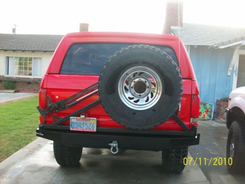 1985 Ford Bronco - The rear view of a red SUV shows a spare tire mounted on a custom bumper, with a jack secured to the side.
