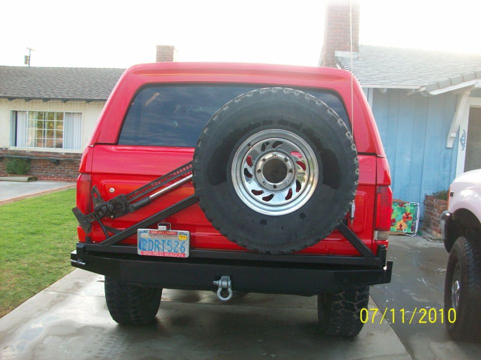 Red SUV's rear bumper with a mounted spare tire and a Hi-Lift jack attached to the side. Another vehicle is partially visible on the right