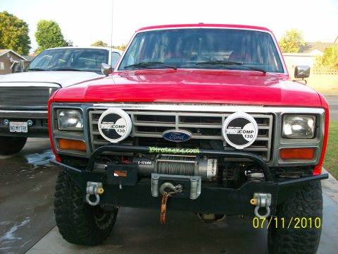 1985 Ford Bronco - Front view of a red Ford Bronco with a winch and auxiliary lights, parked next to a white truck.