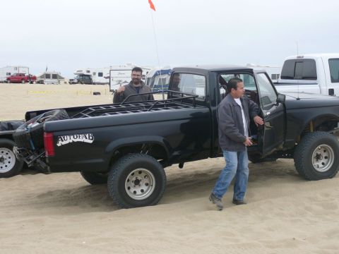 1988 Ford Bronco - Two men stand by a black pickup truck with a custom bed rack on a sandy beach, with RVs and other vehicles in the background.