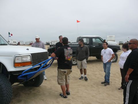 1988 Ford Bronco - A group of people gather around a white Ford F-Series truck with a custom bumper on a sandy terrain, with another pickup truck in the backg…