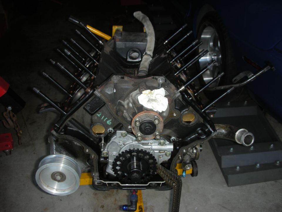 Engine block on stand, showing timing chain, oil pump, and pushrods. A silver pulley is on the left