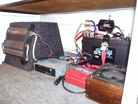 2006 Ford E350 - Auxiliary power and audio setup laid out under a plywood platform: a V-Power amplifier on a carpeted board, a loose head unit, a red 350-wa…