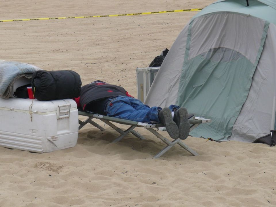 Person sleeping on a cot next to a cooler and a tent on a sandy surface