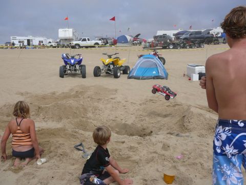 1988 Ford Bronco - A remote controlled car flies through the air on a sandy beach, with children and adults watching.