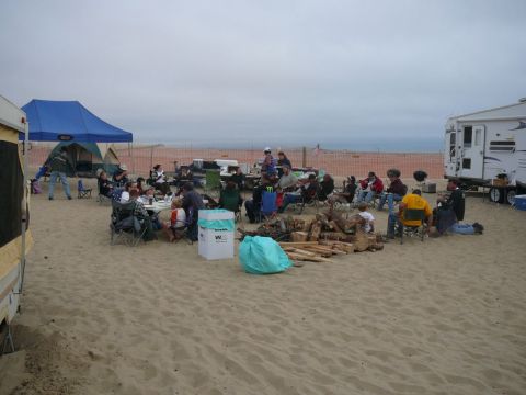 1988 Ford Bronco - A group of people enjoy a beach gathering with RVs and tents set up, gathered around a fire pit on the sand.
