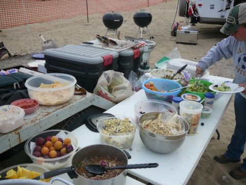 1988 Ford Bronco - A beach picnic features a spread of food, grills, and a person serving salad from a table.
