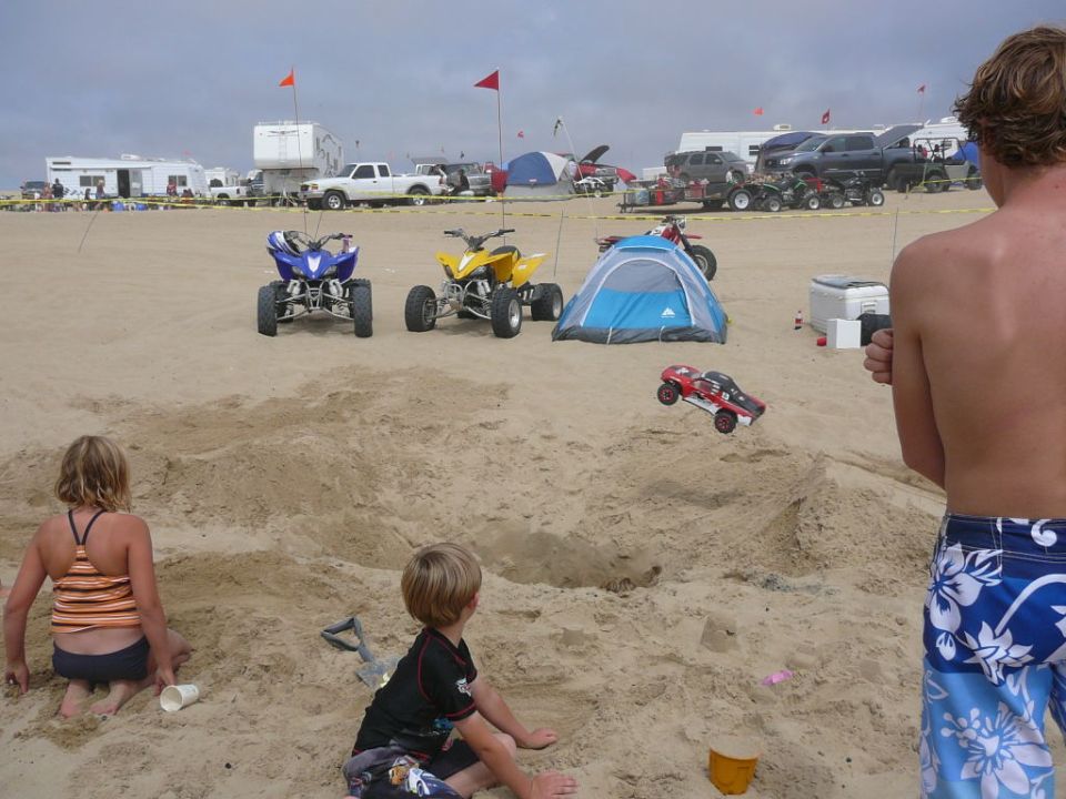Kid playing with remote control car on beach, other kids and ATVs in background