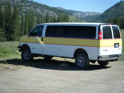 1981 Ford E350 - A white and yellow passenger van is parked on a dirt road with a mountainous, forested background.