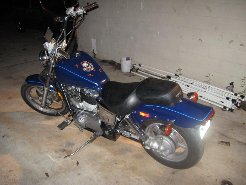 Blue Honda cruiser motorcycle with skull tank graphic, exhaust removed, parked at night by a cinder-block wall