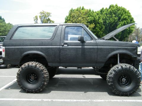 1982 Ford Bronco - This modified SUV features oversized tires and a textured black finish, with its hood raised.