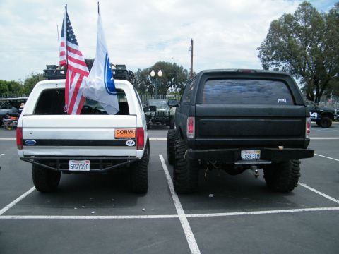 1982 Ford Bronco - Two lifted Ford Broncos are parked in a lot, one white and one black, displaying flags.