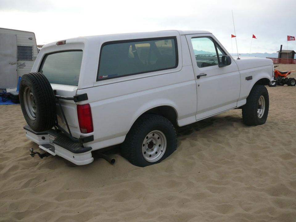 White Ford Bronco stuck in sand, spare tire on back. Trailer and ATV in background