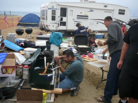 1988 Ford Bronco - A group of people are gathered around a campsite on the beach, cooking and socializing near an RV and tents.