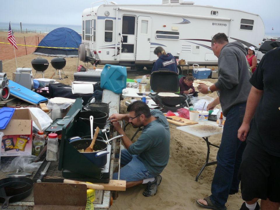 People cooking at a beach campsite with an RV and tents