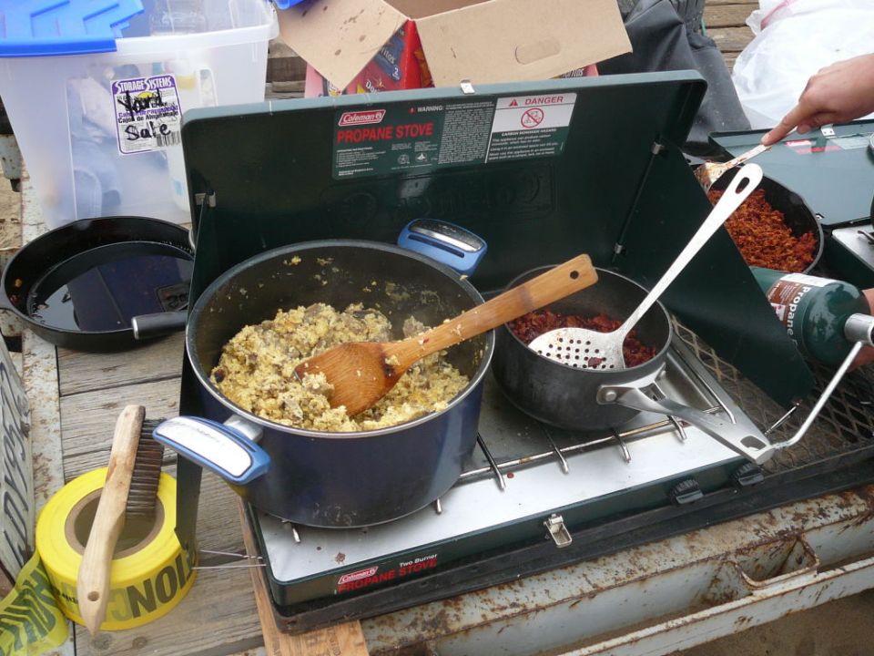 Coleman propane stove with pots of food cooking, wooden spoon, strainer, and propane tank